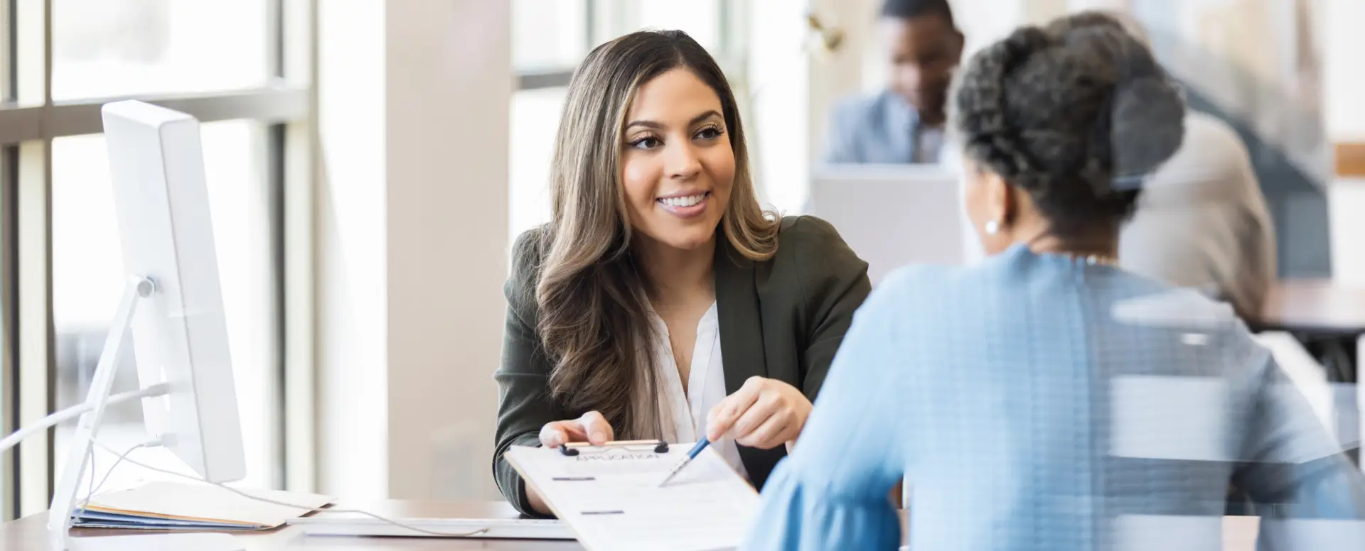 A woman in a blazer smiles and hands a document to a first-time home buyer across a desk in an office setting.