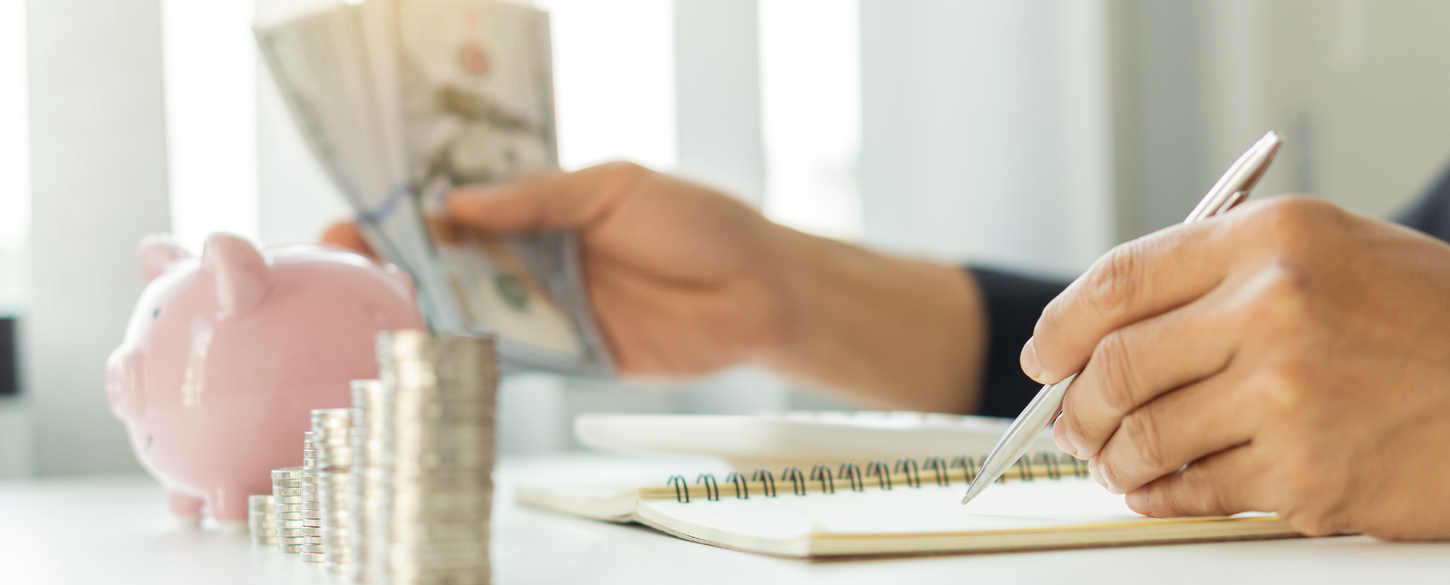 Person counting money, tracking home loans in a notebook, with stacked coins and a pink piggy bank on the table.