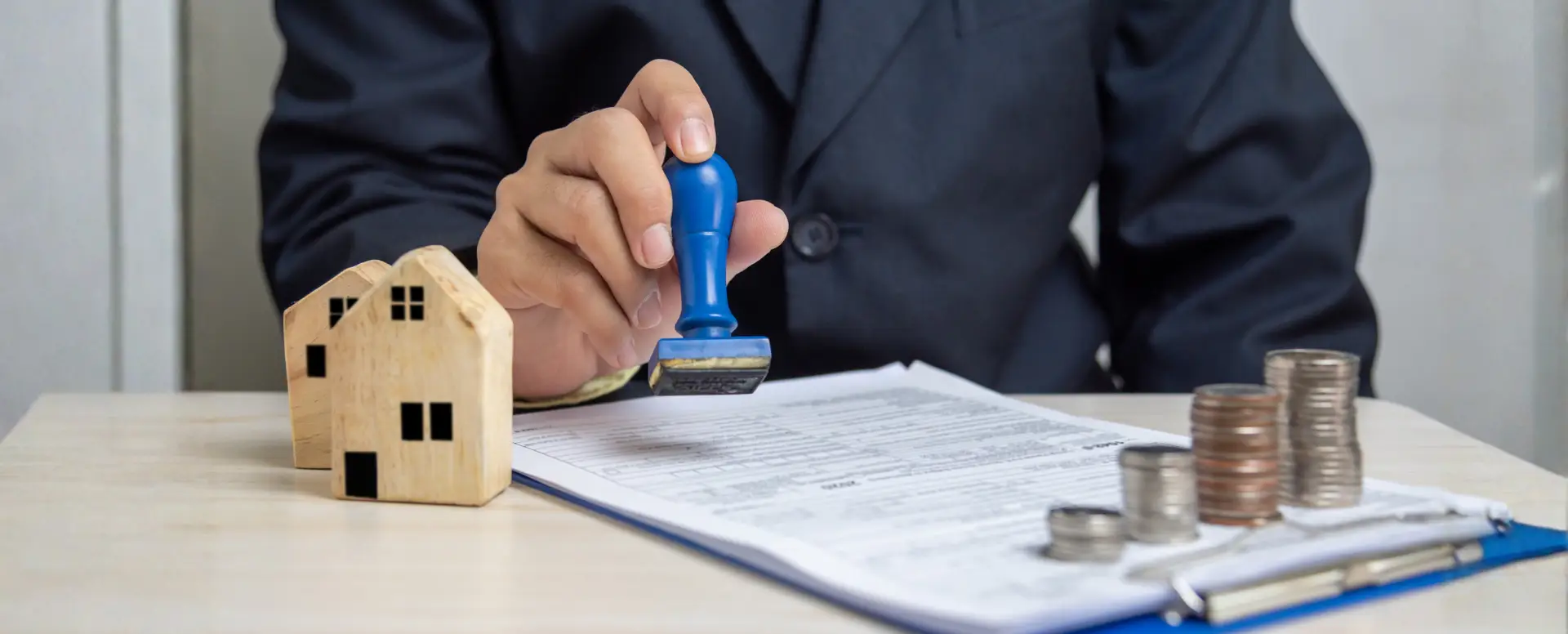 A mortgage lender stamps documents at a desk, with model houses and stacked coins beside them.