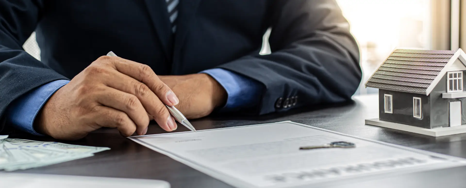 A person in a suit signs a document beside a small house model and key, representing home loans.