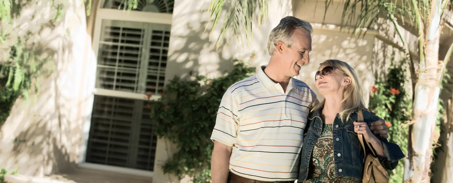 Older couple smiling and embracing outdoors, happy with their mortgage lender in front of a sunlit house and garden.