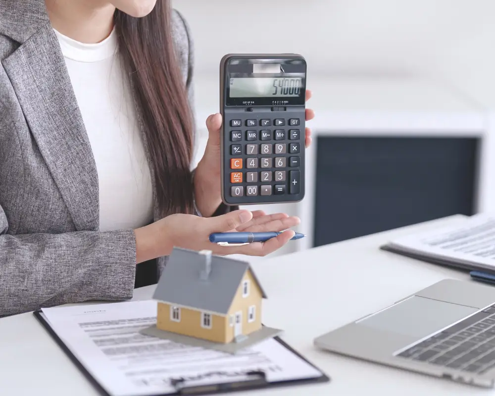 A mortgage lender in a suit holds a calculator showing $50,000 next to a model house and paperwork on a desk.