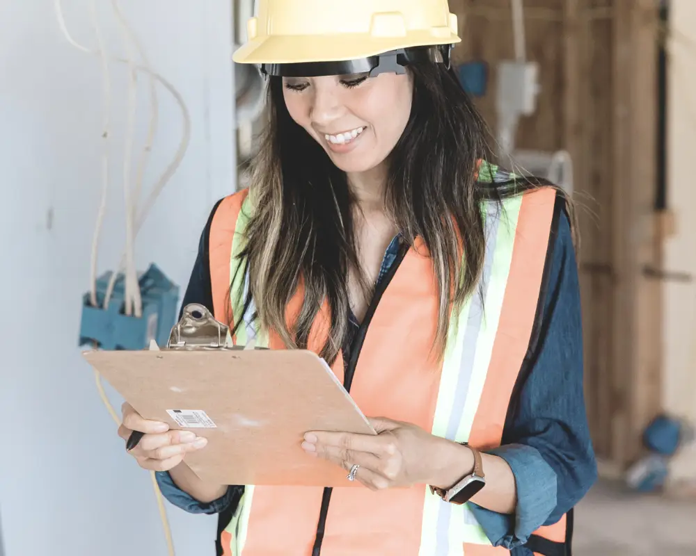 Woman in a hard hat smiles while reviewing construction loans on her clipboard at the building site.
