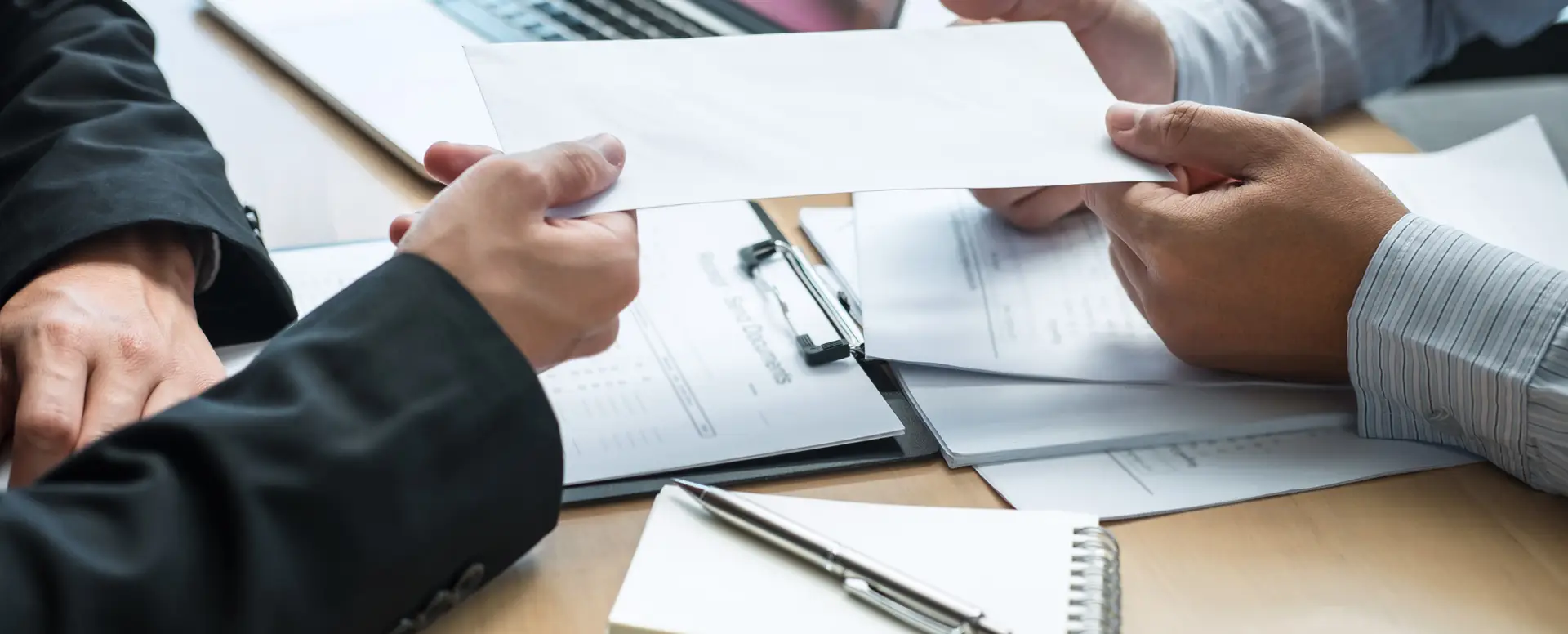 Two people in business attire exchange a mortgage refinance envelope over a desk with documents and a pen.