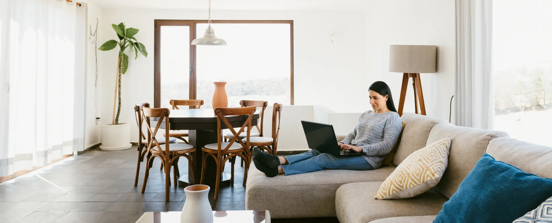 Woman sitting on a beige sofa using a laptop in a bright room, researching home loans in natural light.