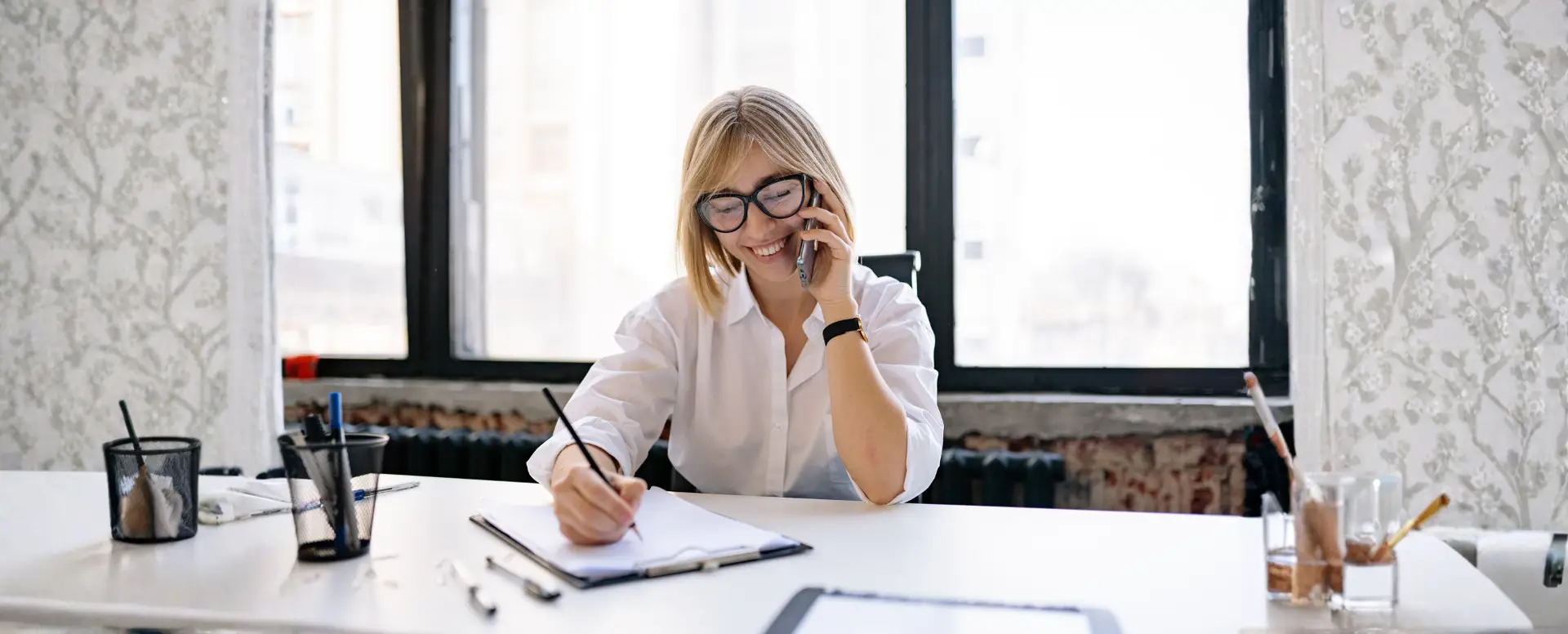 Smiling woman in glasses writes in a notebook while discussing purchase home loans at a bright office desk.