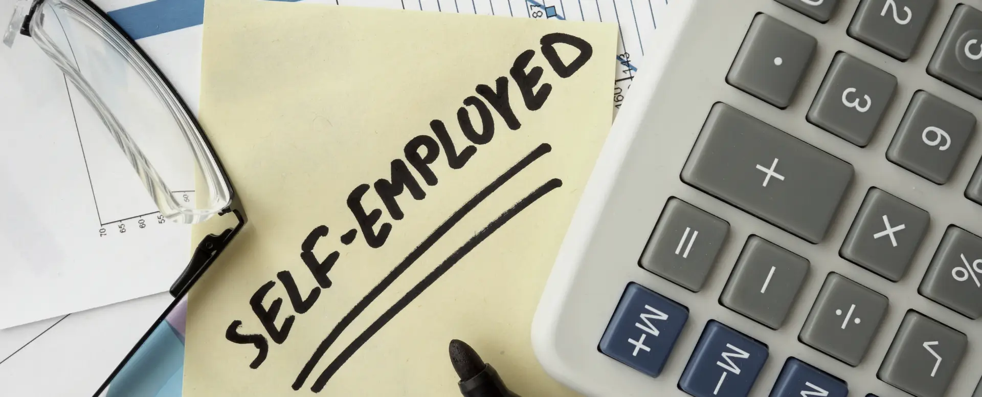 A sticky note with SELF-EMPLOYED written on it, next to a calculator—great for first-time home buyers or home loans.