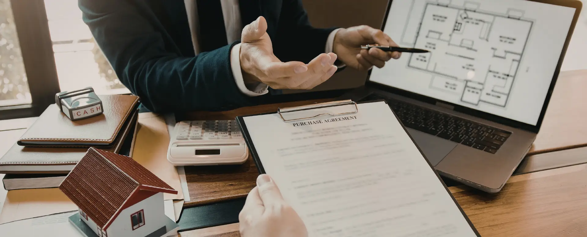 Two people discuss a purchase agreement, with a mortgage lender's floor plan and house model on a laptop in the background.