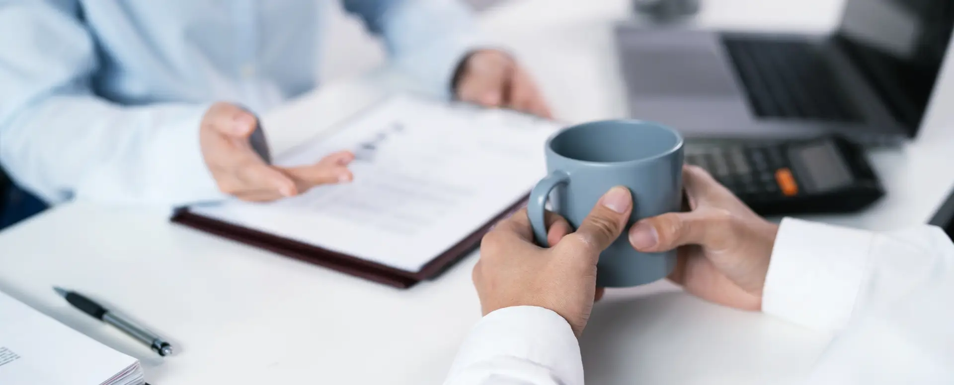 Two people sit at a desk; one holds a coffee mug while the other discusses purchase home loans near a laptop.