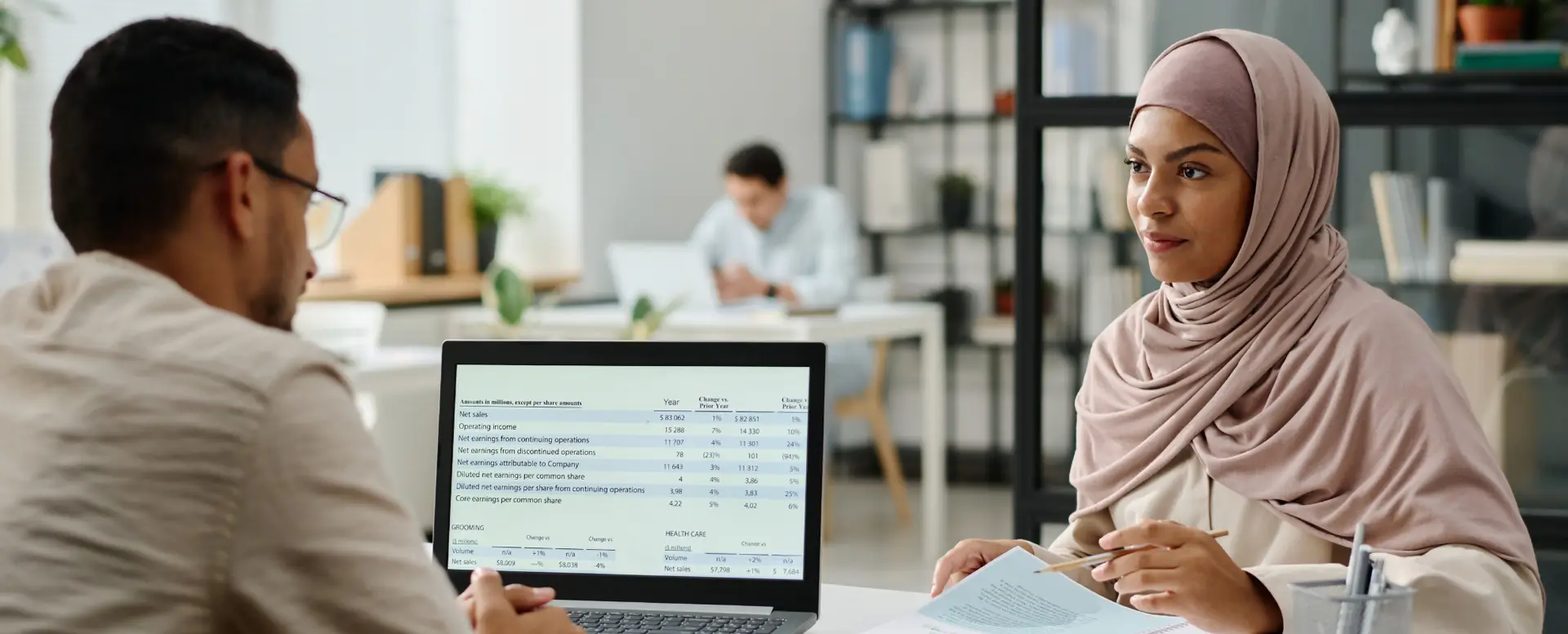Two professionals discuss mortgage refinance data on a laptop, while another person works in the background.