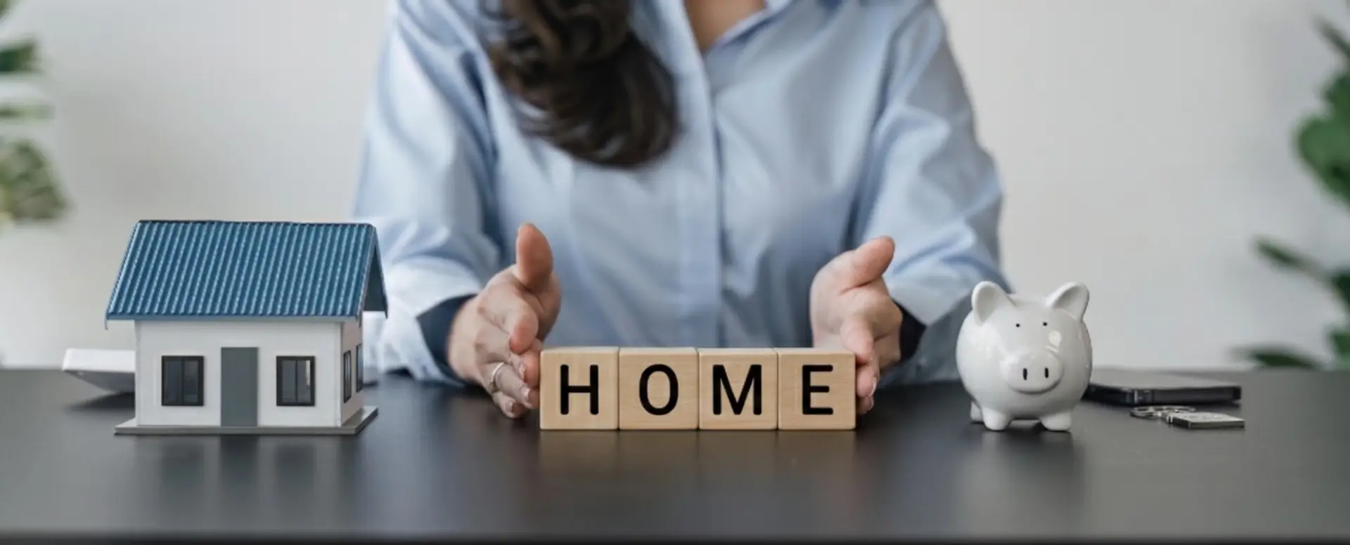 A person sits at a table with a toy house, blocks spelling HOME, and a piggy bank—perfect for first-time home buyers.