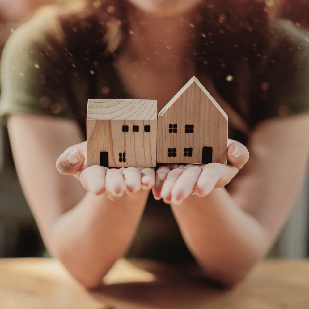 A person holds two small wooden house models, symbolizing home loans for first-time home buyers against a warm background.