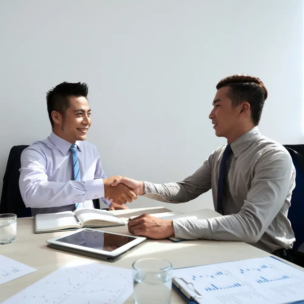 Two businessmen in shirts and ties smiling, shaking hands—discussing purchase home loans at a desk with documents and tablets.