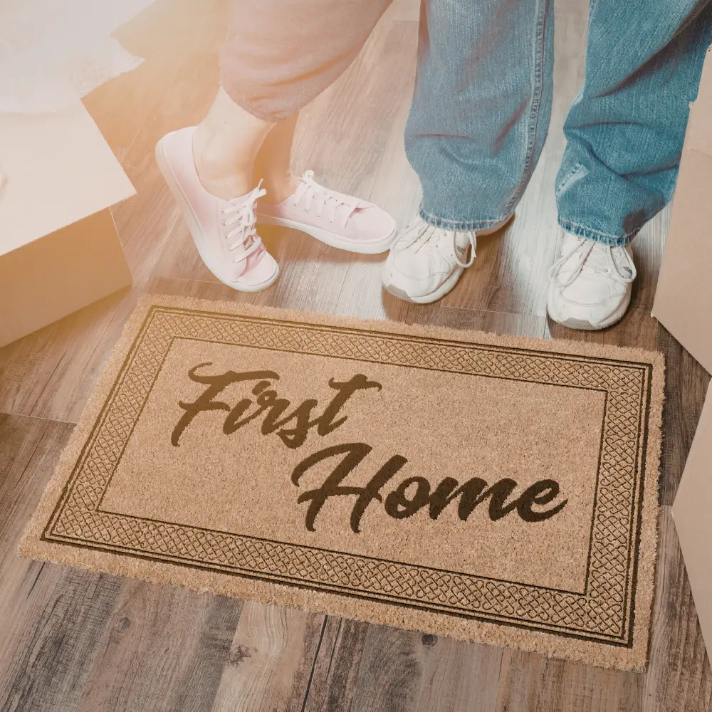 A doormat reading First Home welcomes first-time home buyers, with two people and moving boxes on a wooden floor.