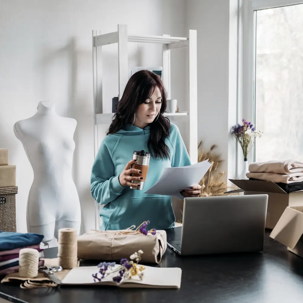 Woman in blue hoodie holding papers and coffee, standing by laptop—researching home loans for first-time home buyers.