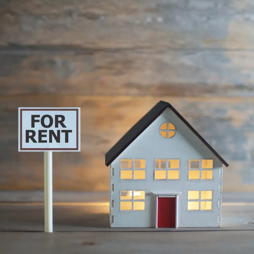 A small model house with lit windows sits by a FOR RENT sign—ideal for first-time home buyers exploring options.